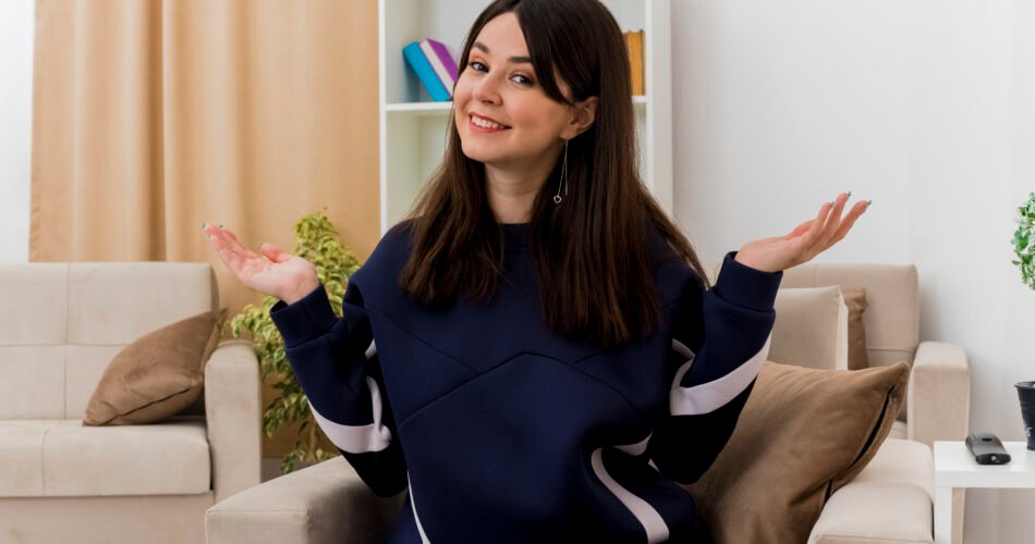 Smiling Young Pretty Caucasian Girl Sitting On Armchair In Designed Living Room Looking At Camera Showing Empty Hands